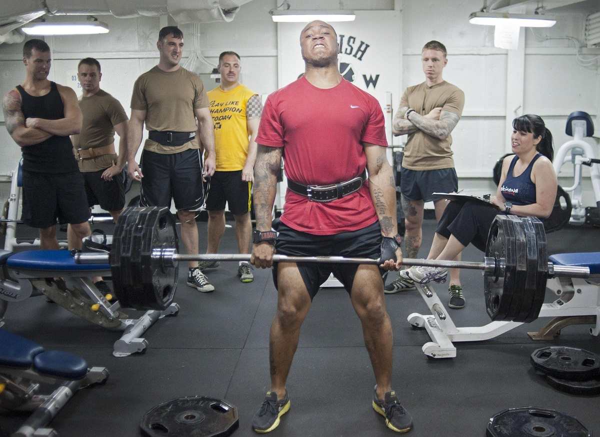 Homme musclé soulevant une barre chargée de poids, entouré de spectateurs dans une salle de musculation Homme musclé soulevant une barre chargée de poids, entouré de spectateurs dans une salle de musculation